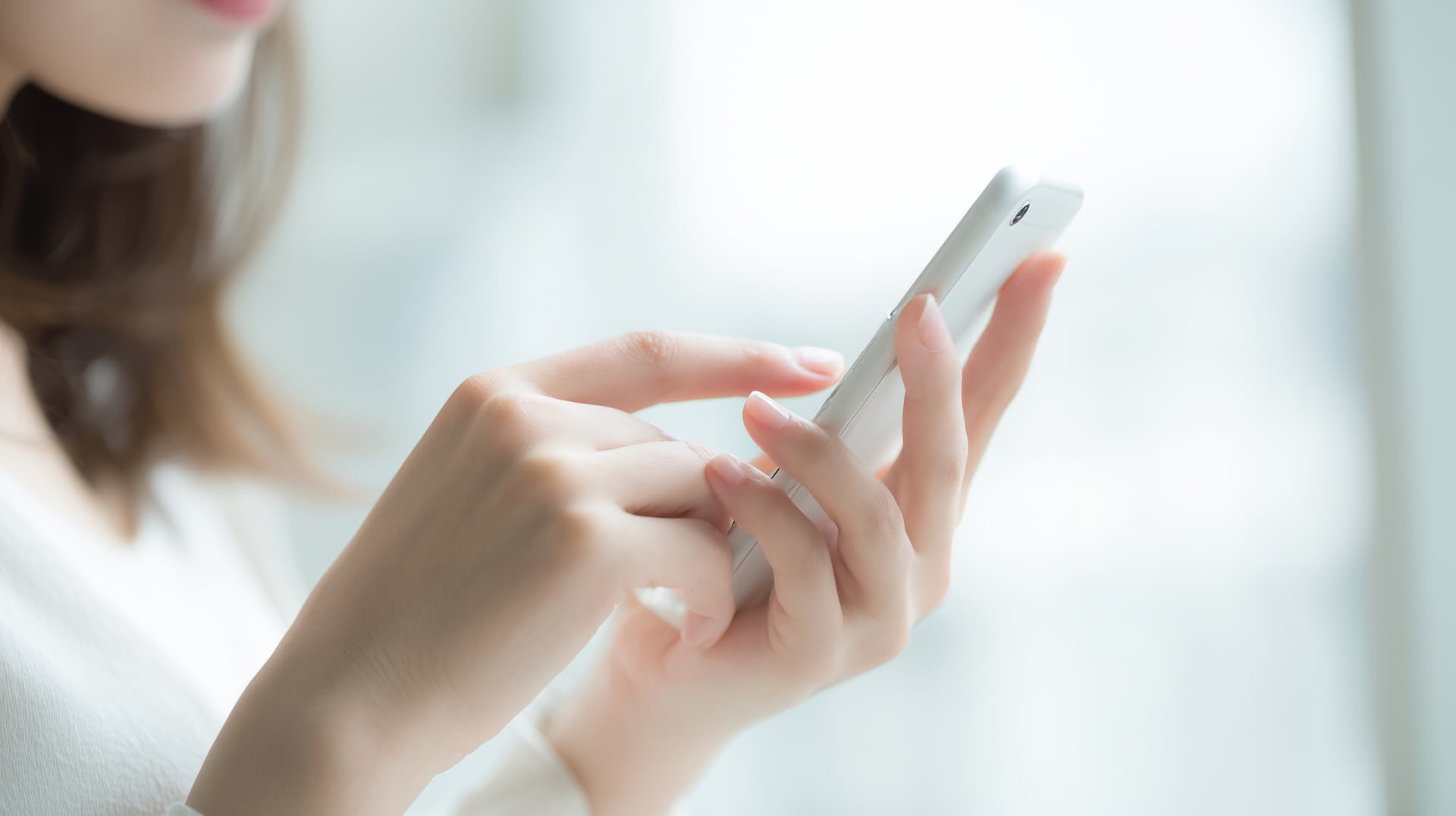 明るい窓の前でスマートフォンをタップする女性の手元